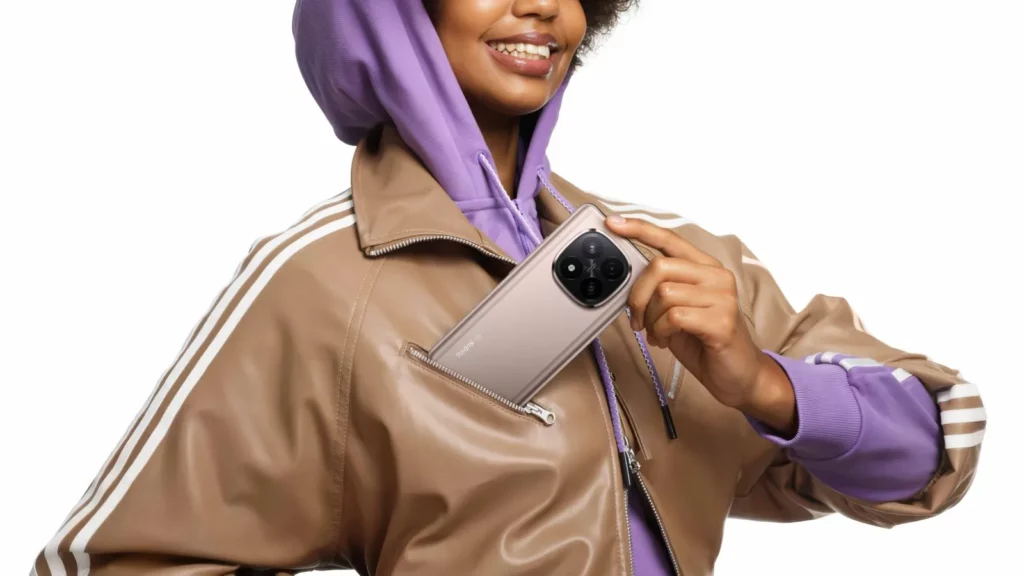 A smiling person in a brown and purple tracksuit jacket and hoodie is pulling a beige or light gold Redmi Note 14 Pro+ 5G smartphone out of their jacket pocket, showcasing the phone's back with its large circular camera module.