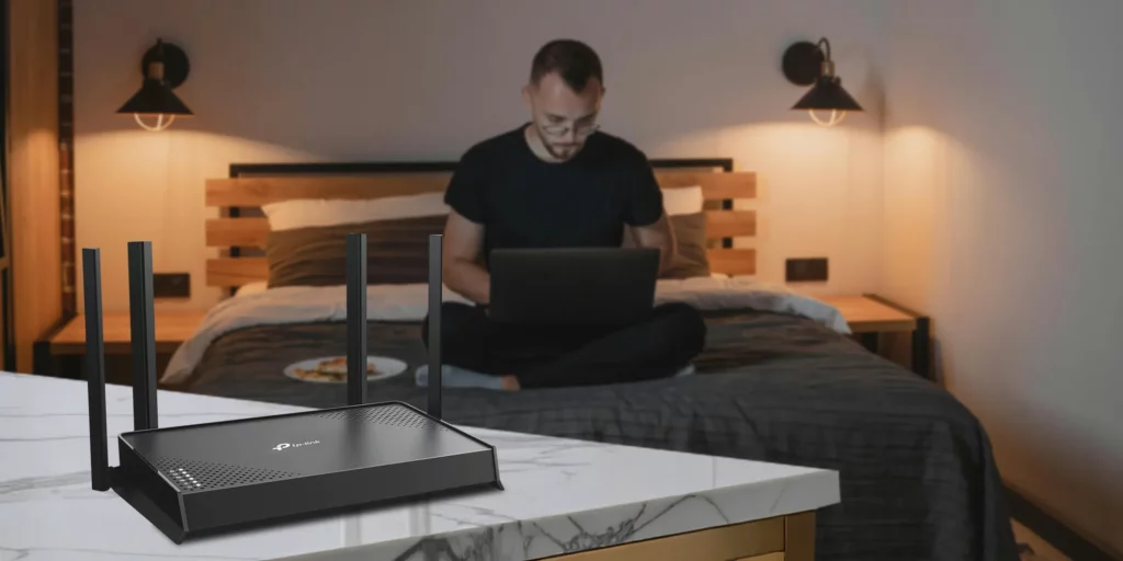 A TP-Link Archer BE220 Wi-Fi 7 router is placed on a kitchen counter in the foreground. In the blurry background, a man sits on a bed with a laptop, suggesting seamless connectivity for working or Browse from any room in the house.