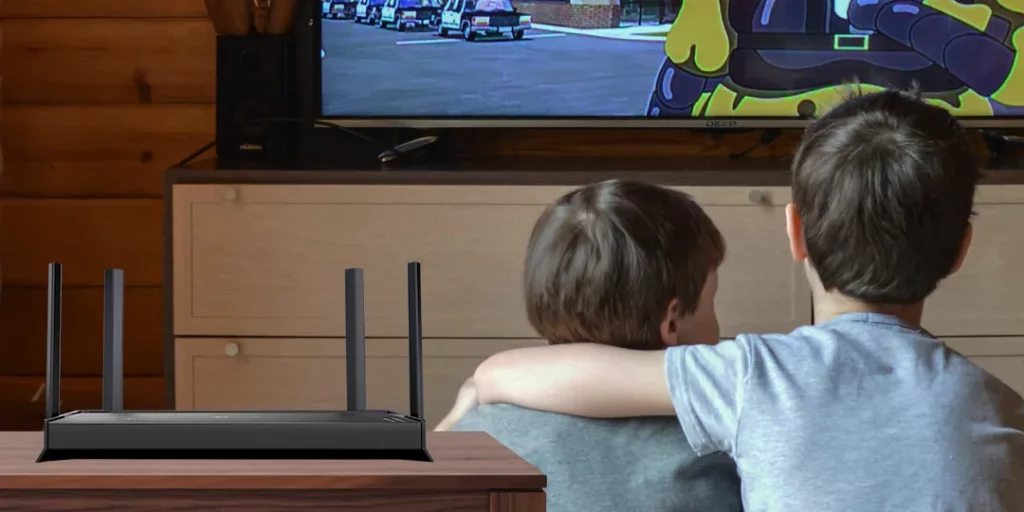 A TP-Link Archer BE220 Wi-Fi 7 router sits on a wooden table in the foreground. In the background, two young children are shown from behind, sitting on a couch and watching an animated show on a television. The scene suggests smooth streaming for entertainment.