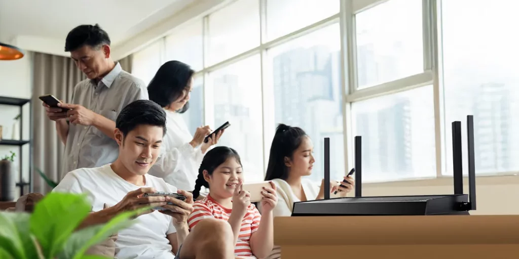 A TP-Link Archer BE220 Wi-Fi 7 router is on a wooden surface in the foreground. A family is gathered in a well-lit living room, with each member—a man, a woman, and two children—using their smartphones or other devices, illustrating multiple devices connected at once.