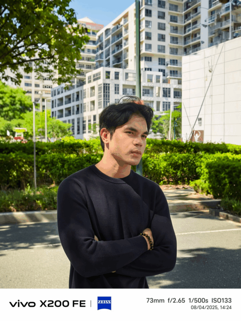 A close-up portrait of a man with his arms crossed, taken outdoors with the vivo X200 FE. The background is slightly blurred, highlighting the camera's bokeh effect. The EXIF data shows the photo was taken at 73mm with a f/2.65 aperture.