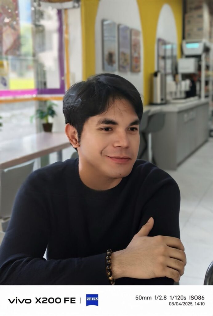 A portrait photo of a man smiling, taken with the vivo X200 FE and its ZEISS camera. The image has a soft focus on the subject, with a blurred background of an indoor cafe. The photo was captured at 50mm, as indicated by the EXIF data.