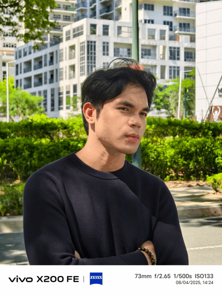 A close-up portrait of a man with his arms crossed, taken outdoors with the vivo X200 FE. The background is slightly blurred, highlighting the camera's bokeh effect. The EXIF data shows the photo was taken at 73mm with a f/2.65 aperture.