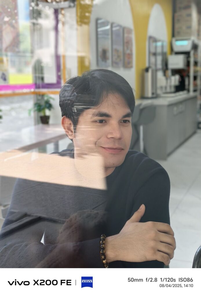 A portrait photo of a man smiling, taken with the vivo X200 FE and its ZEISS camera. The image has a soft focus on the subject, with a blurred background of an indoor cafe. The photo was captured at 50mm, as indicated by the EXIF data.