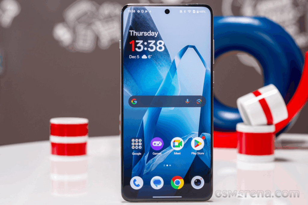 A OnePlus 15 smartphone is standing upright on a white table. The phone's screen is on, displaying a home screen with a digital clock, app icons, and a blue abstract wallpaper. In the background are red, white, and blue cylindrical objects. The GSMArena.com logo is visible in the bottom right corner.