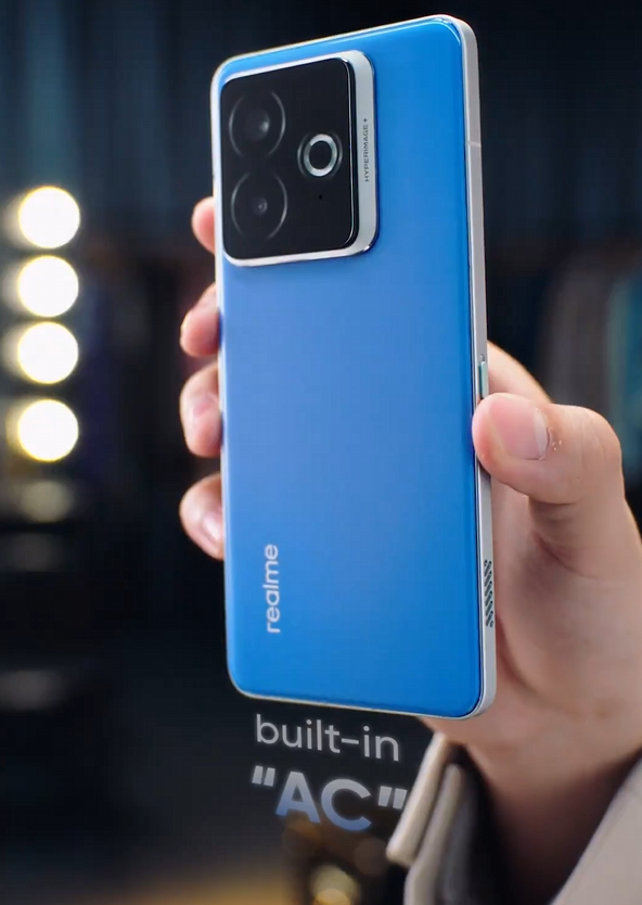 A promotional photo of a hand holding a blue Chill Fan Phone. The phone is shown from the back, highlighting its unique design. It has a flat, light blue back panel with the "realme" logo in white. The camera module is a large, black, asymmetrical rectangle with two circular lenses. A cutout in the center of the camera module reveals a small circular element, possibly a cooling fan. The phrase "built-in 'AC'" is visible in the background, referring to the phone's cooling technology. The text below the image reads "realme Chill Fan Phone via, Gizmochina."