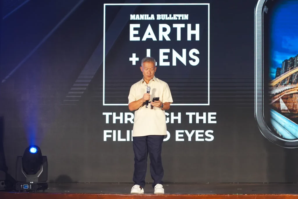 A man standing at a podium, speaking into a microphone while holding a phone. The background features the Manila Bulletin Earth+Lens logo and the theme "Through the Filipino Eyes."
