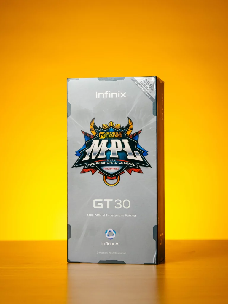 A vertical photo of the Infinix GT 30 retail box in a silver-gray color against a bright orange-yellow background. The box features the Infinix logo, the Mobile Legends: Bang Bang Professional League (MPL) Philippines logo, and text that identifies it as the MPL Official Smartphone Partner. A small sticker on top indicates "256GB + 16GB RAM (8GB Extended)."