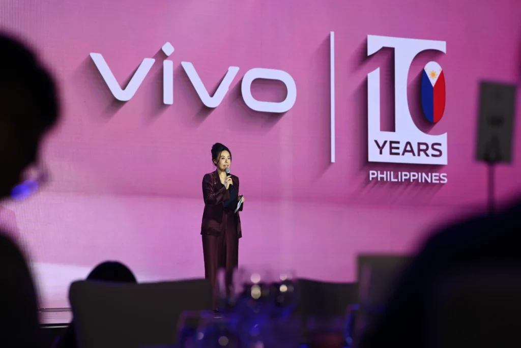 Lulu Liu, Brand Marketing Director for vivo Philippines, stands on a stage giving an opening speech. Behind her is a large screen with the "vivo" logo and "10 YEARS PHILIPPINES" text. The lighting is pink, and the foreground is blurred, focusing on her.