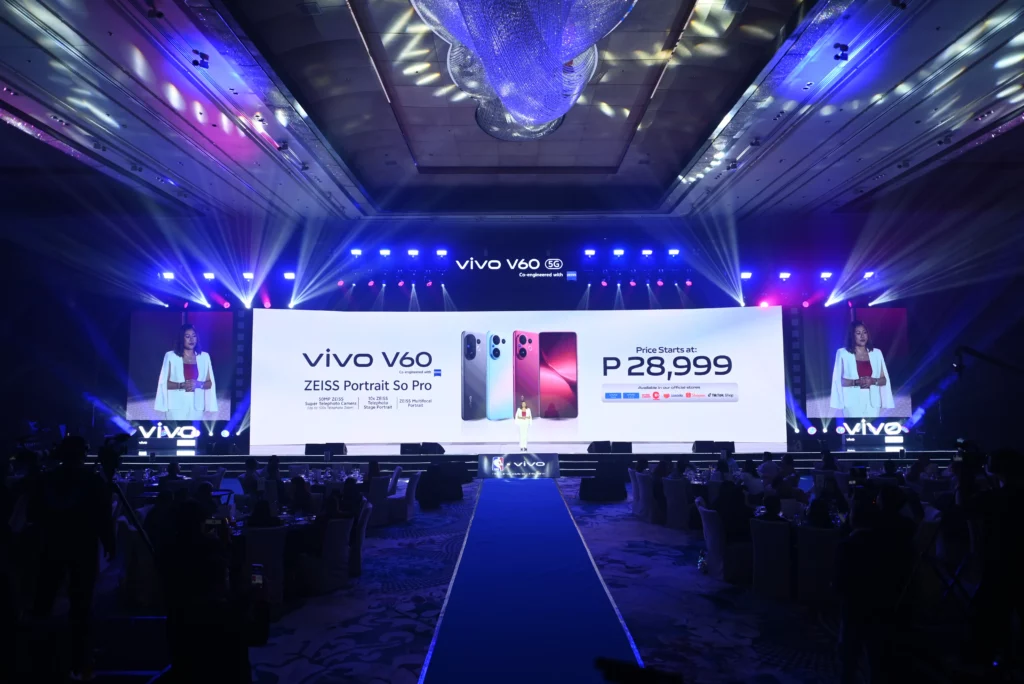 A wide shot of a large, well-lit stage at the vivo V60 launch event. A speaker stands in the middle of the stage, with a long blue carpet leading to them. A massive screen behind them displays the "vivo V60" logo, product images of the phones, and the official price reveal: "Price Starts at: P 28,999."