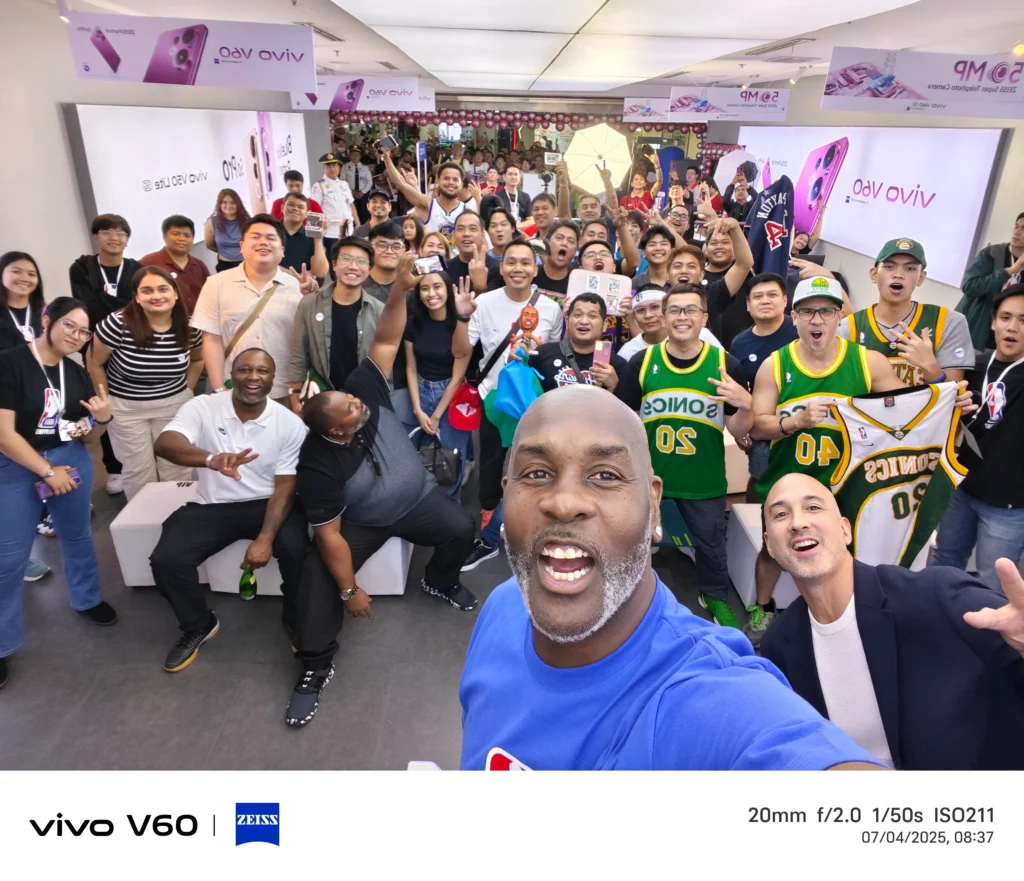 A close-up group selfie taken by NBA icon Gary Payton, showing him and a diverse crowd of fans celebrating at the vivo store event. Payton is smiling, with fans in basketball jerseys and event shirts posing behind him. The image footer shows the "vivo V60 | ZEISS" logo.