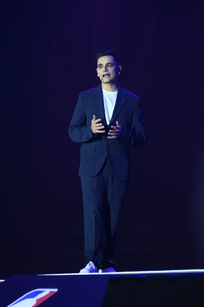 Keshav Chugh, Senior Product Manager at vivo Global, stands on a dark stage with a spotlight on him. He is wearing a dark blue suit over a white t-shirt and is speaking with his hands gesturing outwards.