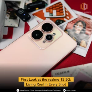 A close-up, top-down shot of the back of a light pink or rose gold Realme 15 5G smartphone. The triple-camera system is the main focus, with three distinct camera lenses and a flash. The phone is placed on a table surrounded by other items, including printed photos and a Hollyland device. The image has a watermark with the text "Unbox Diaries," "Vince Domingo," and "TechBeans," and the title "First Look at the realme 15 5G: Living Real in Every Shot" is at the bottom.