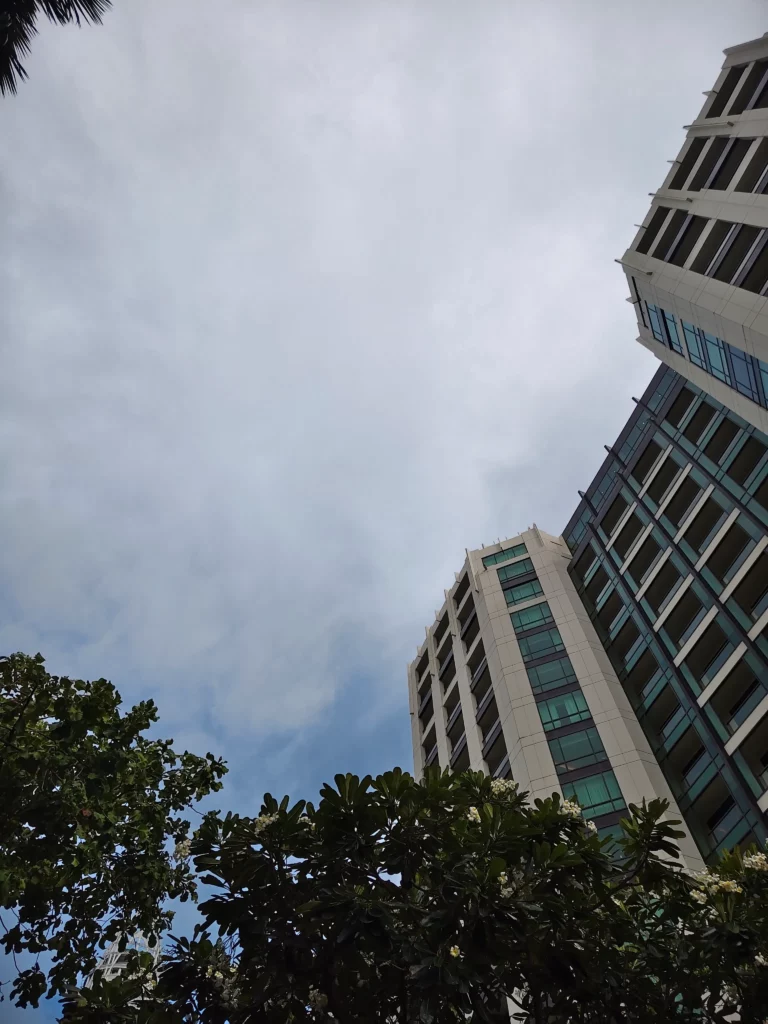 An ultra-wide angle shot captured with the Vivo V60, looking up at two tall buildings and the sky, with trees in the foreground.