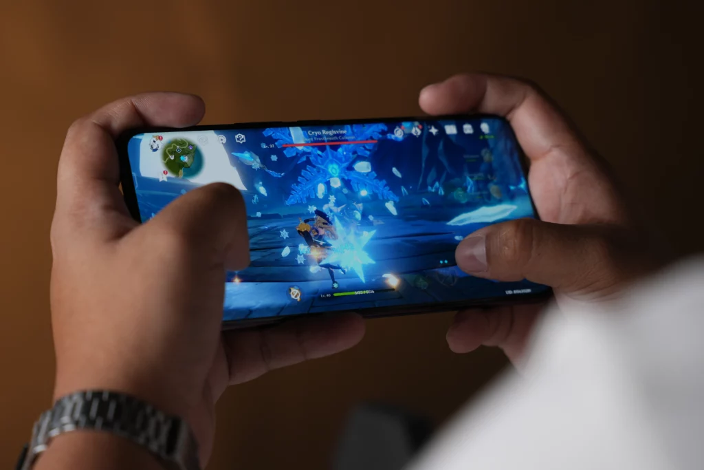 A hands-on shot of a person playing the game Genshin Impact on the Vivo V60, showing an intense in-game battle scene on the phone's screen.