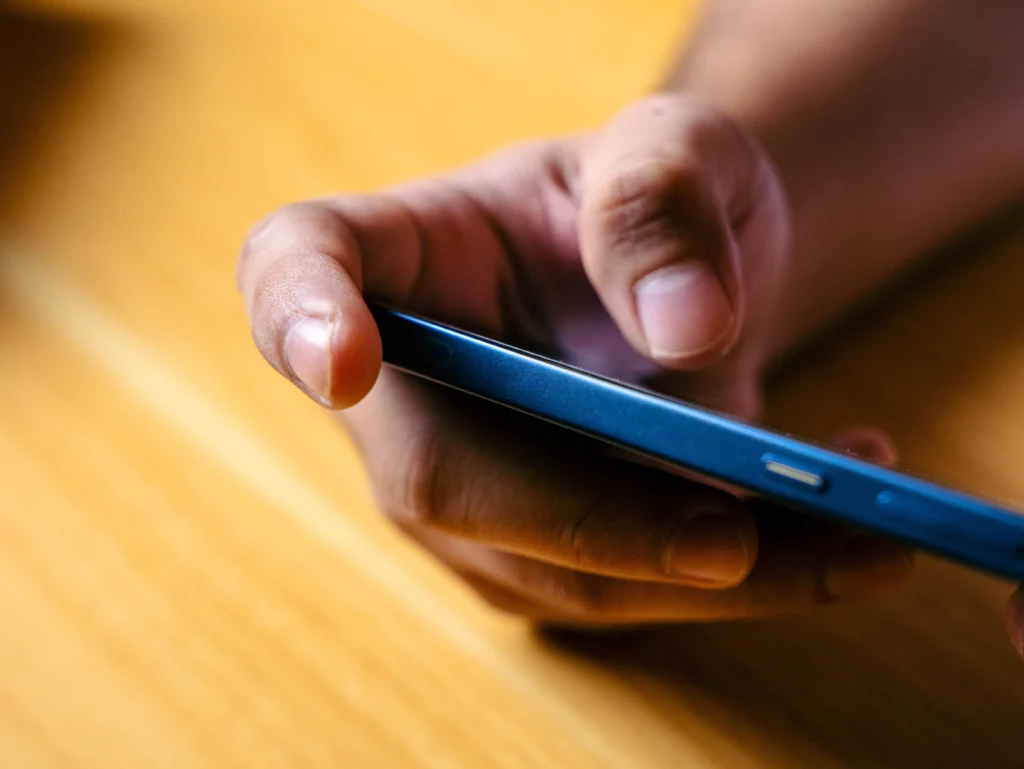 A close-up of a person's hand holding the blue Infinix GT smartphone horizontally over a wooden surface. The focus is on the side profile of the phone, showing the volume rockers and power button, with the thumb and fingers gripping the device.
