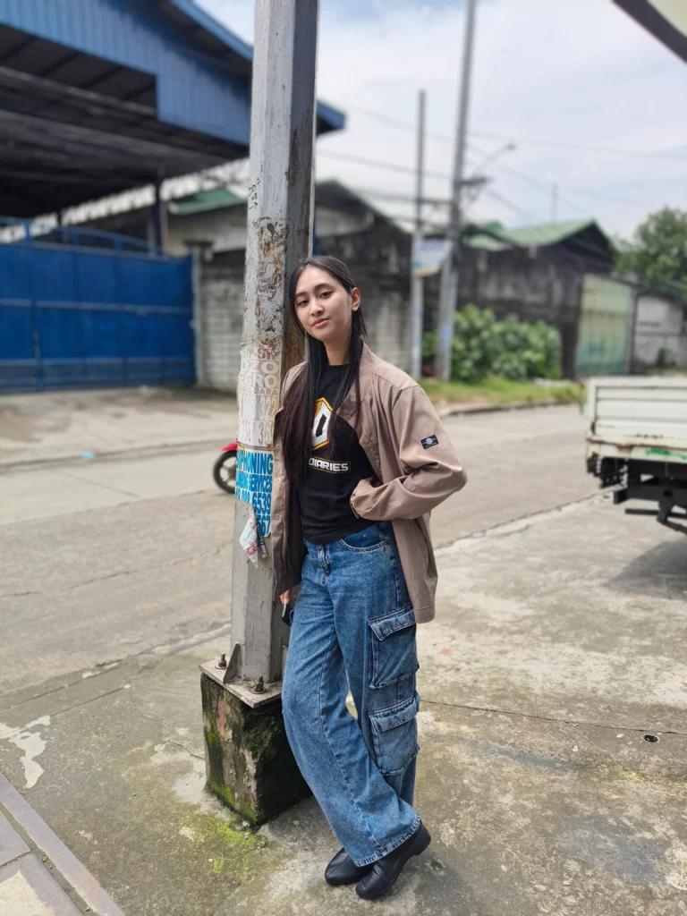 A full-body portrait of a young woman standing on a paved street, leaning against a utility pole. She is wearing a black graphic T-shirt, a beige jacket, and wide-leg denim cargo pants. The photo has a shallow depth of field (bokeh effect), blurring the background of houses and a truck.