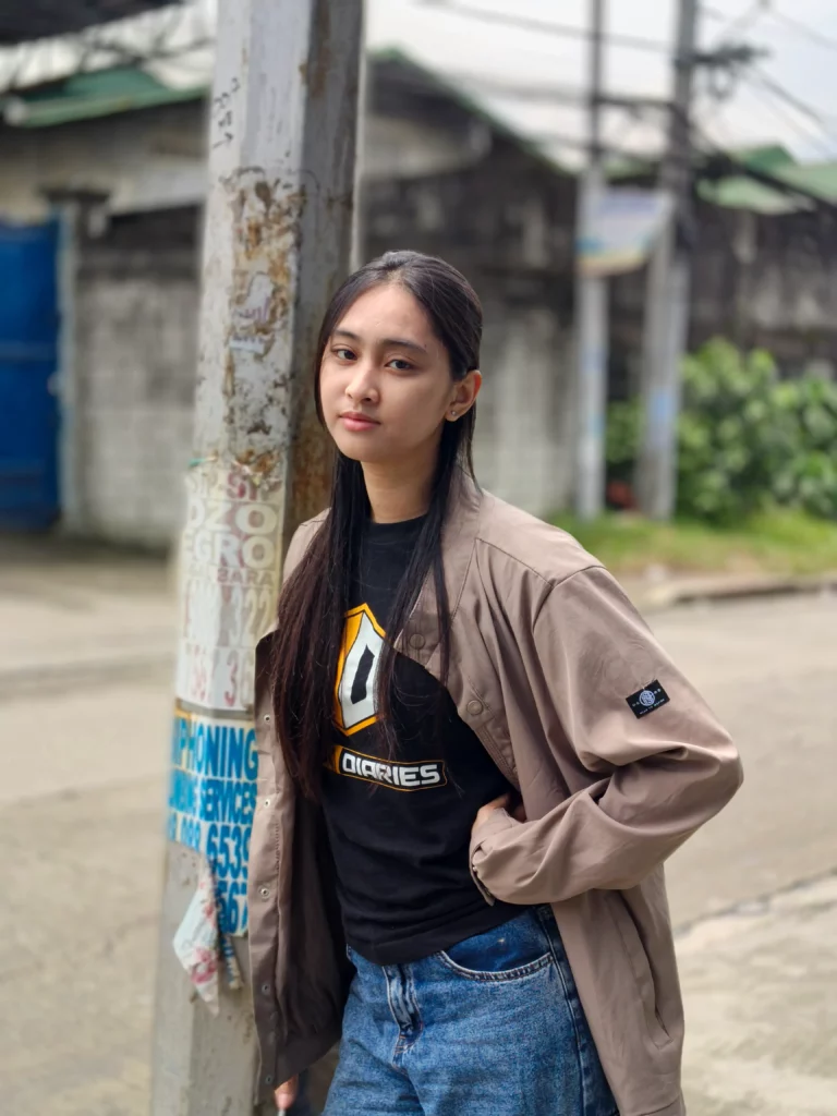 A medium close-up portrait of a young woman standing outdoors, leaning against a utility pole covered in old posters. She has long, dark hair and is wearing a black T-shirt and a beige jacket. The background is slightly blurred.
