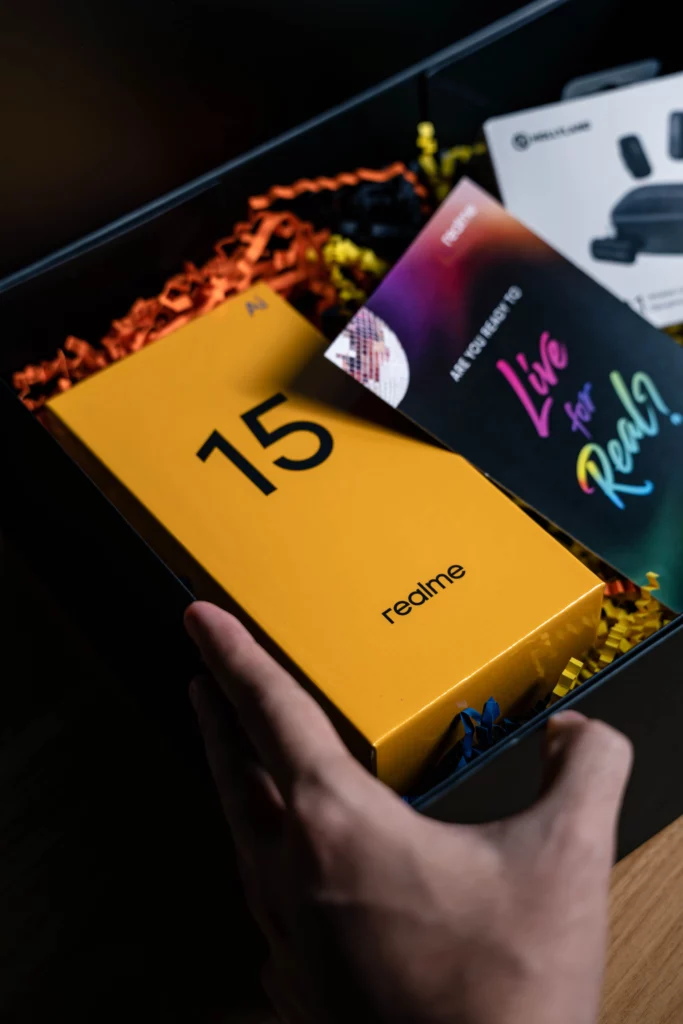 A hand reaching into an open black gift box filled with yellow and orange shredded paper, revealing a yellow realme 15 phone box and a colorful booklet.