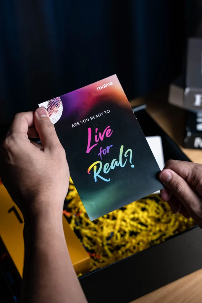 A pair of hands holding a booklet that reads "realme - ARE YOU READY TO Live for Real?" The booklet has a colorful, psychedelic design. A gift box with yellow shredded paper is visible in the background.