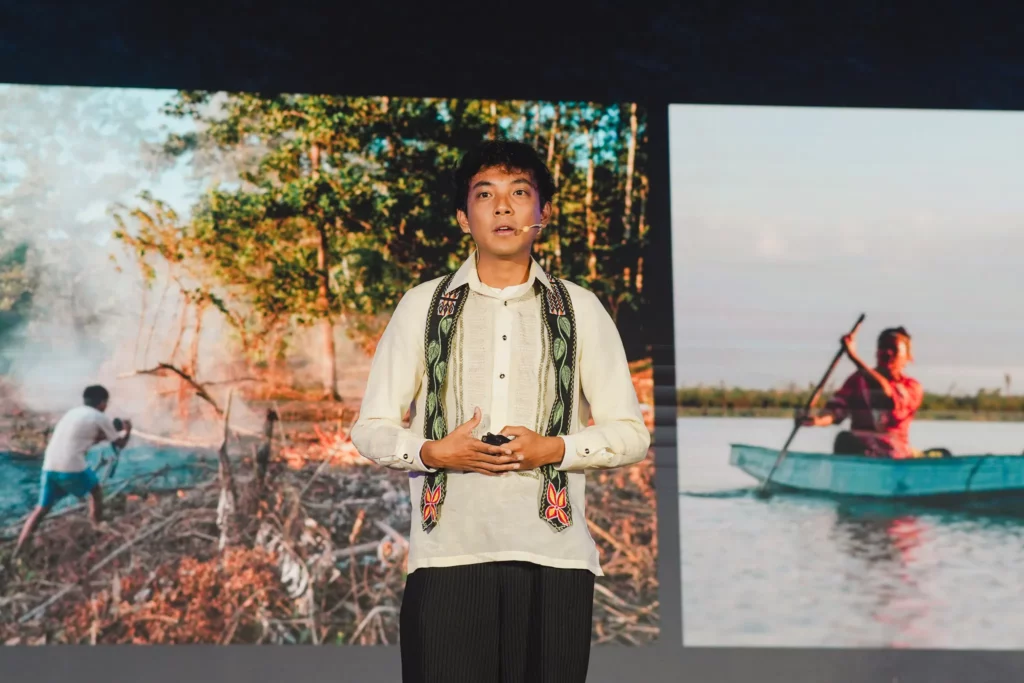 National Geographic Explorer Gab Mejia speaking on a stage. Behind him, two large screens display photographs from the Earth+Lens campaign, one showing a person photographing a burning landscape and the other a person paddling a boat.