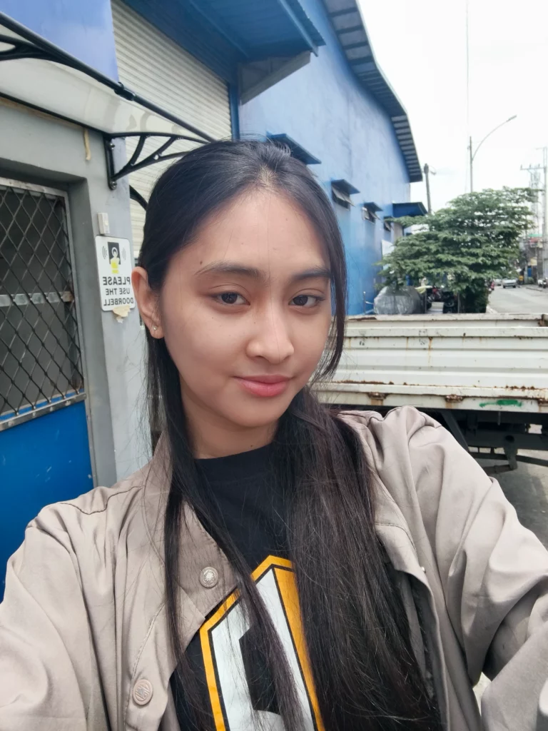 A selfie (front-camera portrait) of a young woman standing outdoors. She is wearing a black graphic shirt and a beige jacket. The background is a street scene featuring a blue warehouse building and the back of a small white truck.