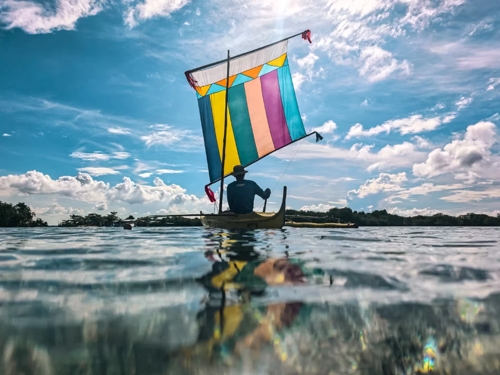 A low-angle shot of a man in a small boat with a vibrant, multicolored sail, gliding across a calm body of water under a bright, partly cloudy sky. The waterline is visible in the foreground.