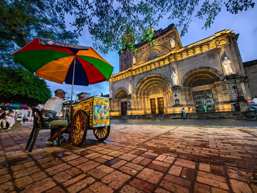 A man selling street food from a pushcart with a colorful umbrella. He is sitting on a bench in a brick plaza in front of the Manila Cathedral at dusk.