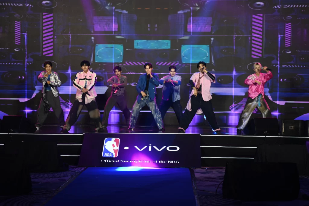 The boy group HORI7ON performs a dance routine on a stage. They are dressed in casual, stylish outfits with colorful, bold patterns. The stage is lit with pink and blue lights, and a large screen behind them displays abstract graphics.