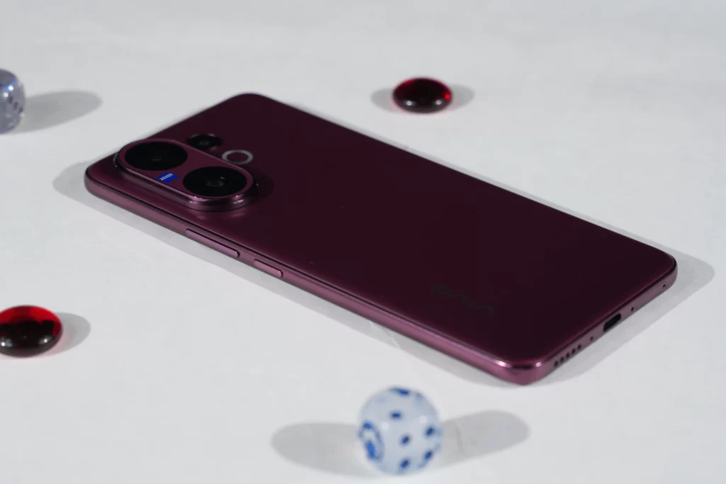 A high-angle, close-up shot of the back of the Vivo V60 smartphone, showcasing the shiny, deep purple finish and the camera module, with the phone resting on a white surface with small decorative glass stones.