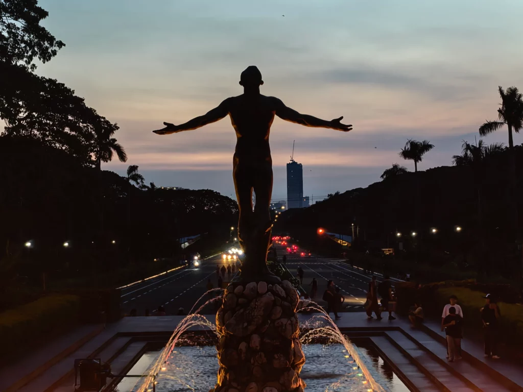 A silhouette of the Oblation statue at the University of the Philippines Diliman, standing in front of a fountain with light trails from cars on a street behind it during twilight.