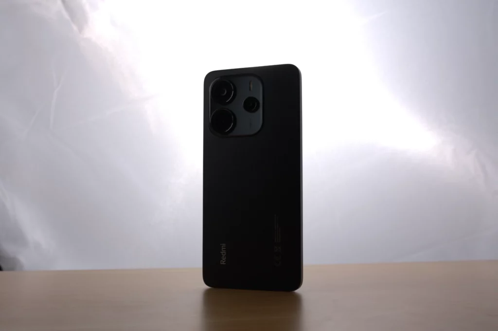 A black Redmi Note 14 smartphone stands upright on a wooden table, with its camera module facing the viewer, against a bright white background.