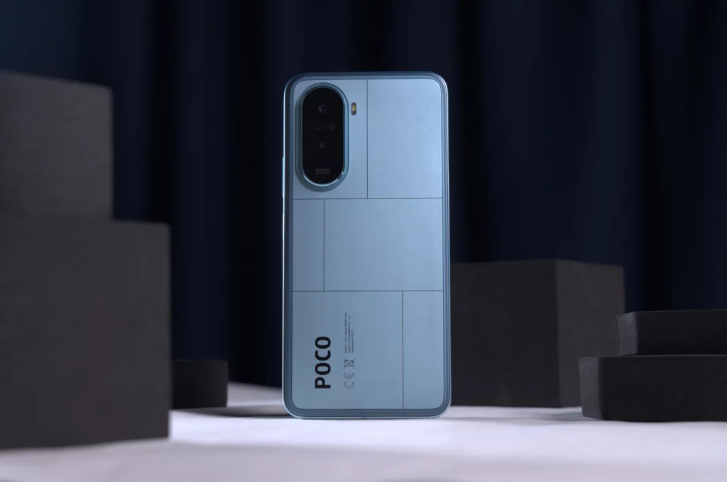A light blue POCO smartphone with a geometric pattern on the back stands upright, surrounded by dark gray foam blocks.