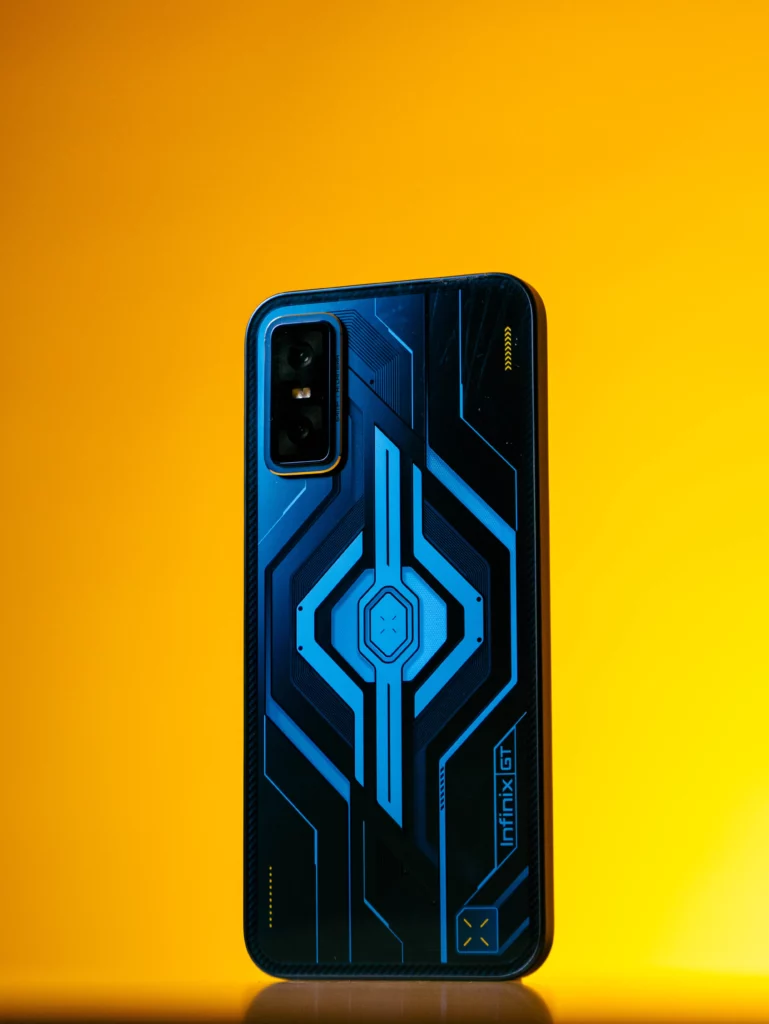 A full vertical shot of the Infinix GT 30 smartphone's back panel. The vibrant Cyber Blue design shows off a central, glowing blue hexagon and intricate surrounding lines. The "INFINIX GT" logo is near the bottom right, and the phone's camera module is visible in the top left, all illuminated by a strong yellow backlight.