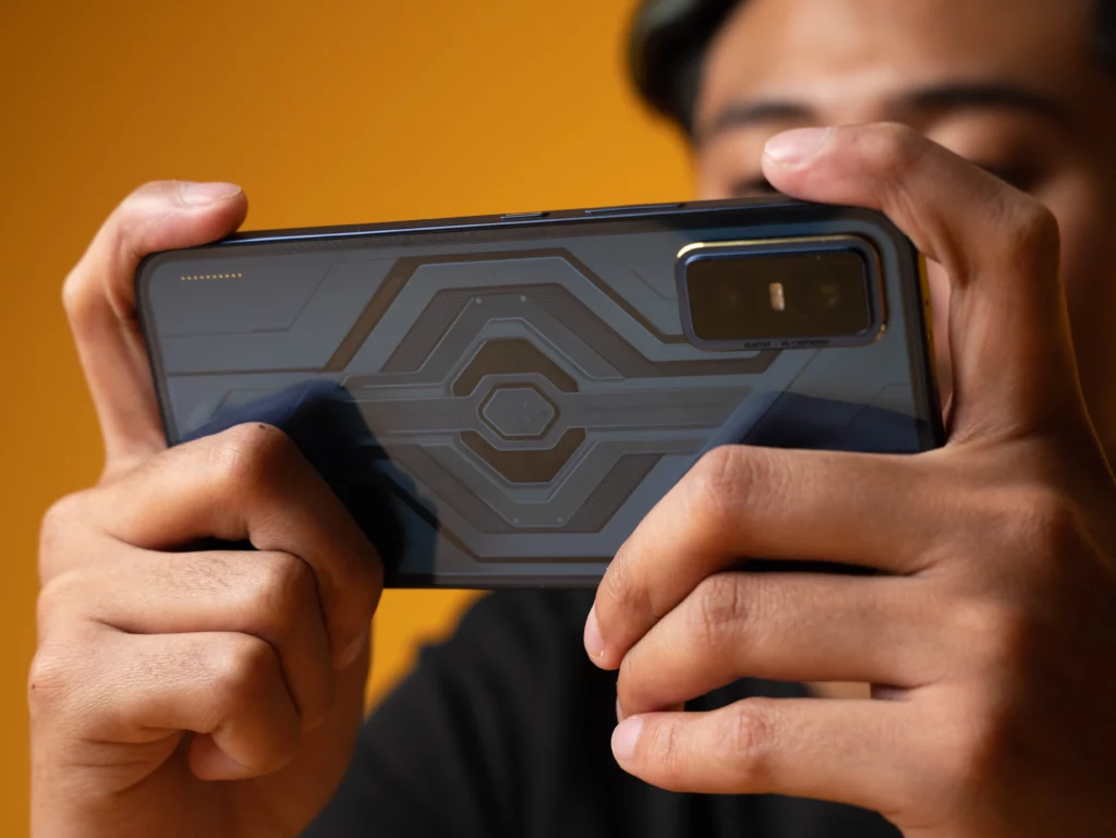 A close-up shot of a person's hands holding the back of the Infinix GT 30 series gaming smartphone horizontally. The phone's back has a dark, geometric design, with the camera module visible on the right.