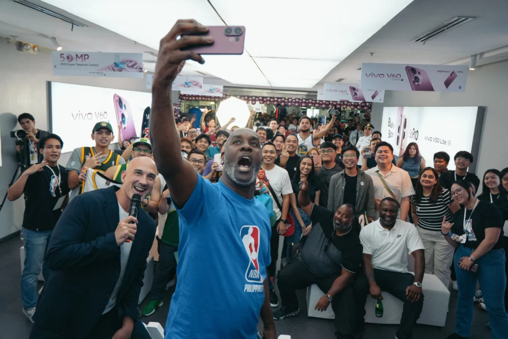 A wide shot of NBA legend Gary Payton taking a group selfie with excited Filipino basketball fans inside the vivo concept store in Manila. Payton is holding a pink vivo V60 smartphone, capturing the crowd in the background.
