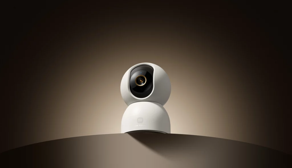 A clean, white, spherical Xiaomi Smart Camera C701 sitting on a subtle brown surface. The camera lens is dark and features a gold ring, facing forward against a dramatic, dimly lit background.