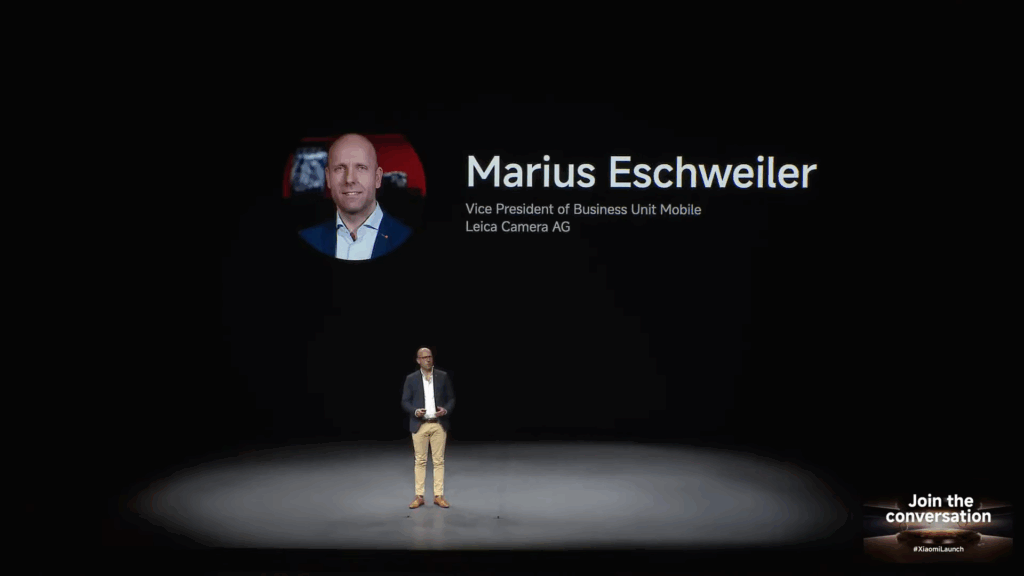 A speaker named Marius Eschweiler is introduced on stage at the launch event. His title is listed as Vice President of Business Unit Mobile, Leica Camera AG.