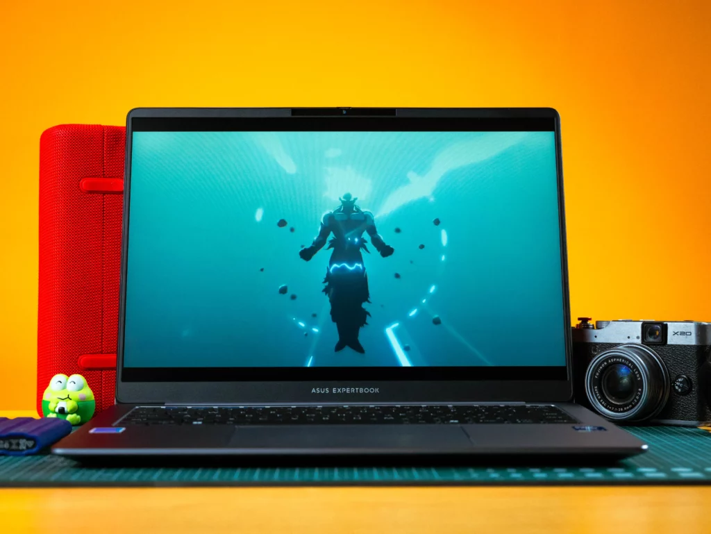 The open ASUS ExpertBook laptop is centered on a desk, displaying a cinematic image of a silhouette of a figure surrounded by bubbles underwater in a teal-colored scene. The laptop is flanked by a red speaker on the left and a silver camera on the right.