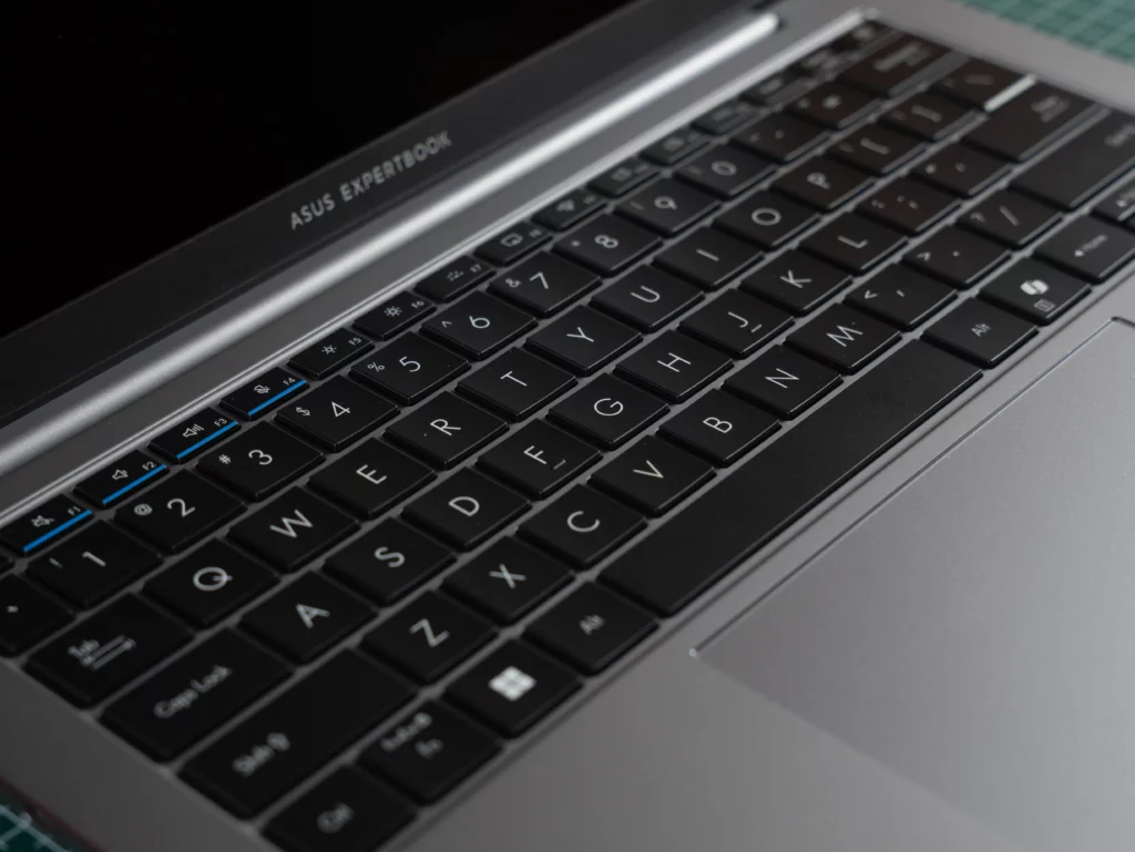 A close-up view of the laptop's backlit keyboard and trackpad. The keys are black, and the function keys (F1-F12) feature blue icons for media and system controls.