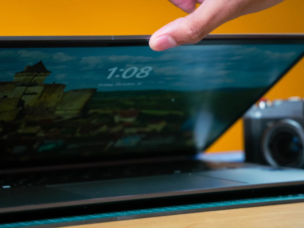 A close-up shot of a hand lifting the lid of the laptop. The top edge of the screen shows the webcam and possibly an infrared camera, with a clock on the screen displaying "1:08."