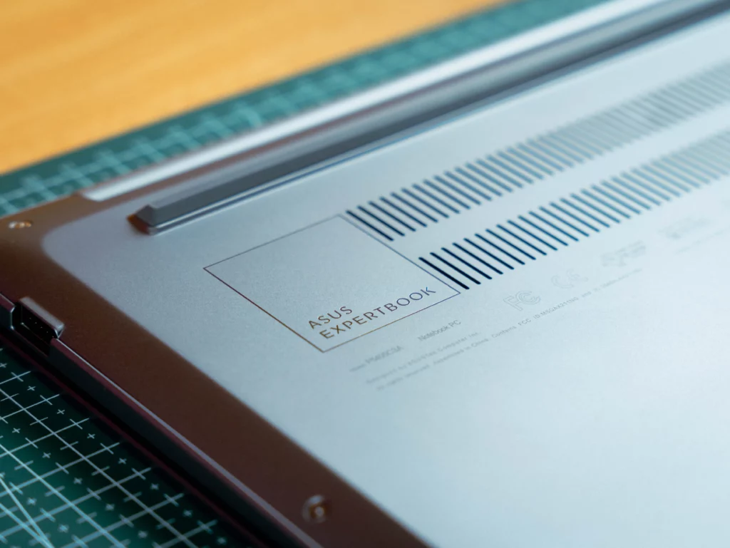 A close-up, angled view of the bottom panel of the laptop, highlighting the engraved "ASUS EXPERTBOOK" logo next to a ventilation grille and various regulatory marks like "FC CE."