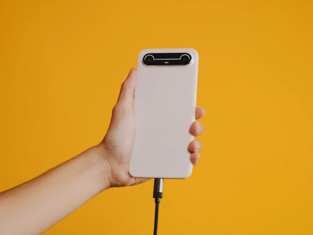 A hand holds the white TECNO SPARK Slim phone vertically, which is connected to a charging cable plugged into the bottom port, against a yellow background.
