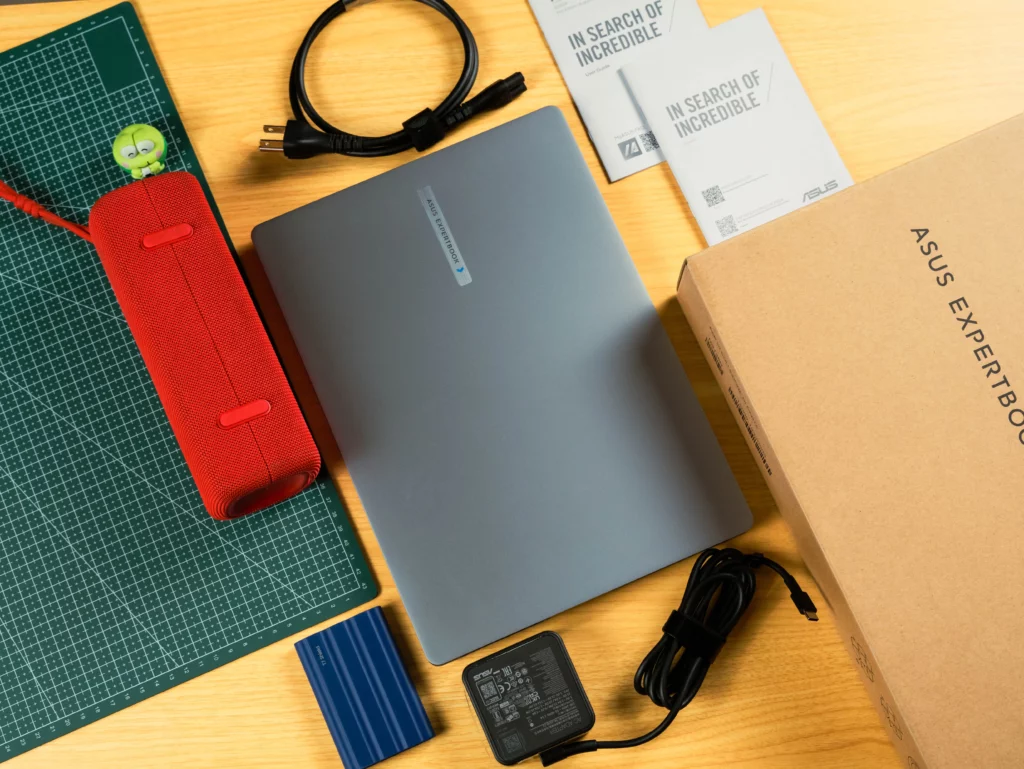 An overhead shot of the ASUS ExpertBook laptop (closed) and its accessories unpacked from the box. Items include the laptop, a power brick and cables, two small paper booklets, a red cylindrical speaker, and a small blue external hard drive, all laid out on a green cutting mat and wooden desk.