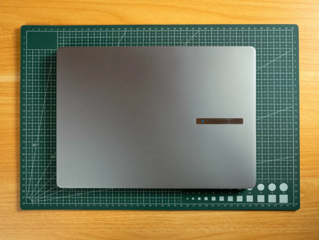 An overhead shot of the closed gray ASUS ExpertBook laptop resting squarely on a green, gridded cutting mat, which is positioned on a wooden desk. A small brown and blue sticker is visible on the laptop's lid.