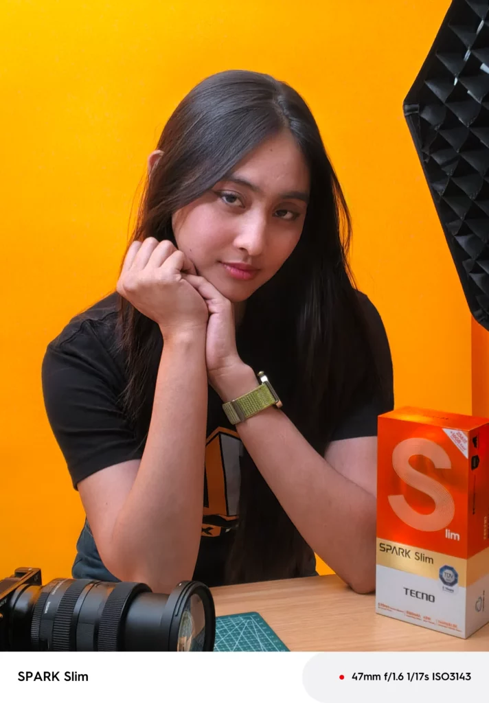 Sample portrait photo taken with the TECNO SPARK Slim, showing a young woman posing next to the phone's orange retail box.