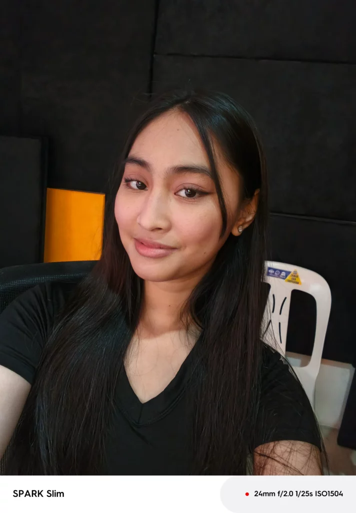 Indoor sample selfie (or front-facing camera photo) taken with the TECNO SPARK Slim of a young woman with long dark hair, smiling at the camera.