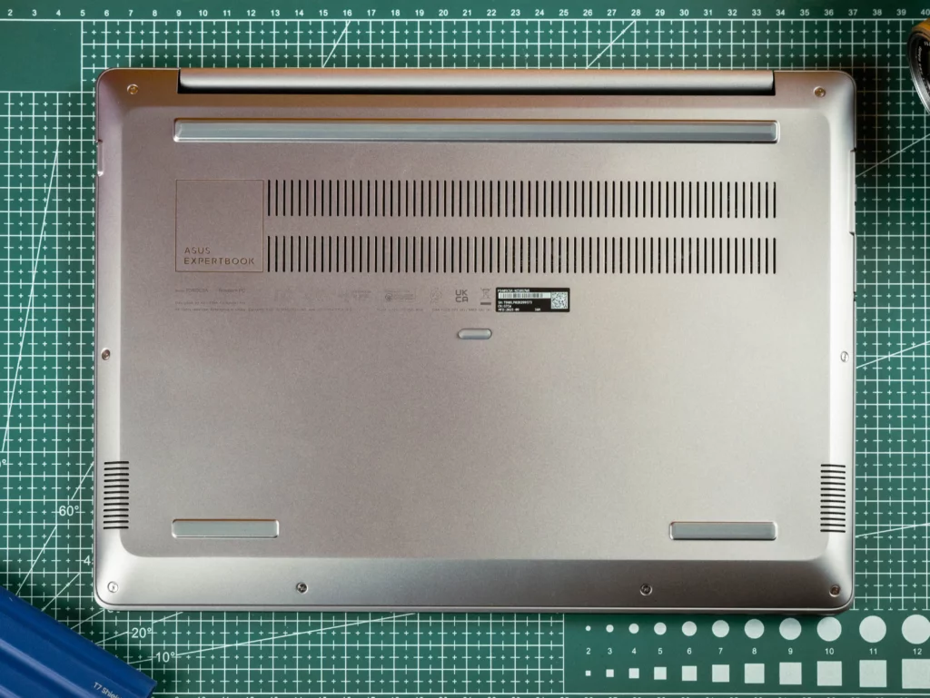 A detailed view of the bottom panel of the ASUS ExpertBook. It is a light metallic gray with two large rows of ventilation grilles in the center, rubber feet, and regulatory/model information stickers in the middle. The laptop is lying on a green cutting mat.