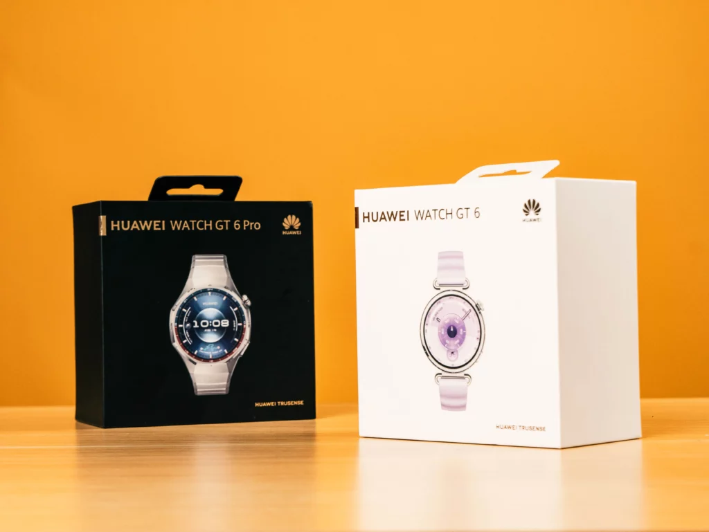Two product boxes on a wooden table against an orange background. The box on the left is black for the Huawei Watch GT 6 Pro, and the box on the right is white for the Huawei Watch GT 6.