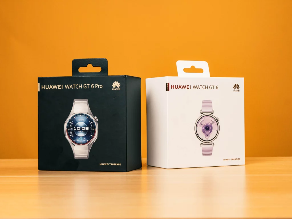 Two rectangular product boxes on a wooden table against an orange background. The box on the left is black and says HUAWEI WATCH GT 6 Pro. The box on the right is white and says HUAWEI WATCH GT 6. Both feature an image of the watch model inside.
